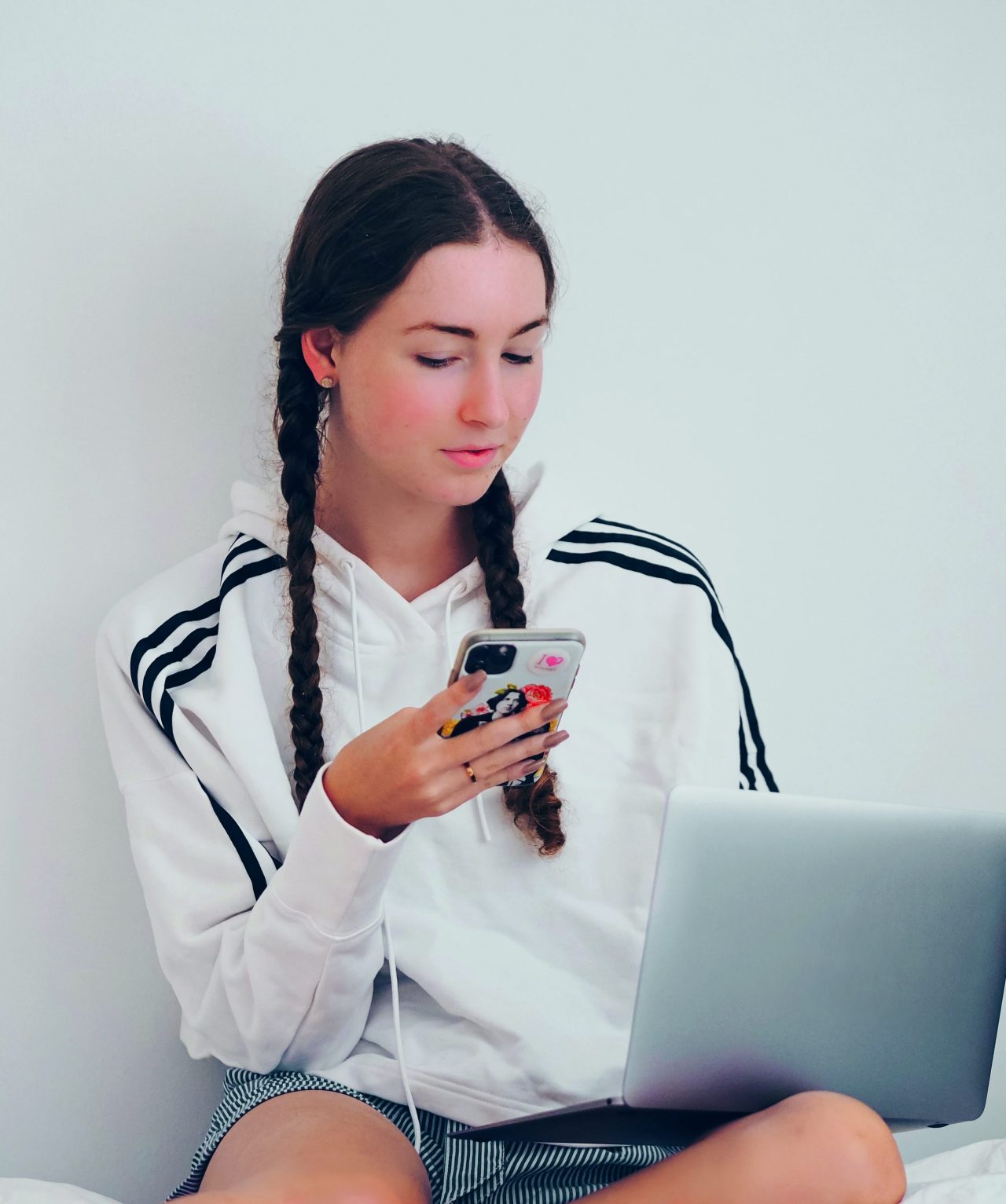 laura-chouette-qaRaOPQbZhI-unsplash Kopie (1) Eine Jugendliche sitzt auf dem Bett und schaut aufs Handy, während sie einen Laptop auf dem Schoß hat.